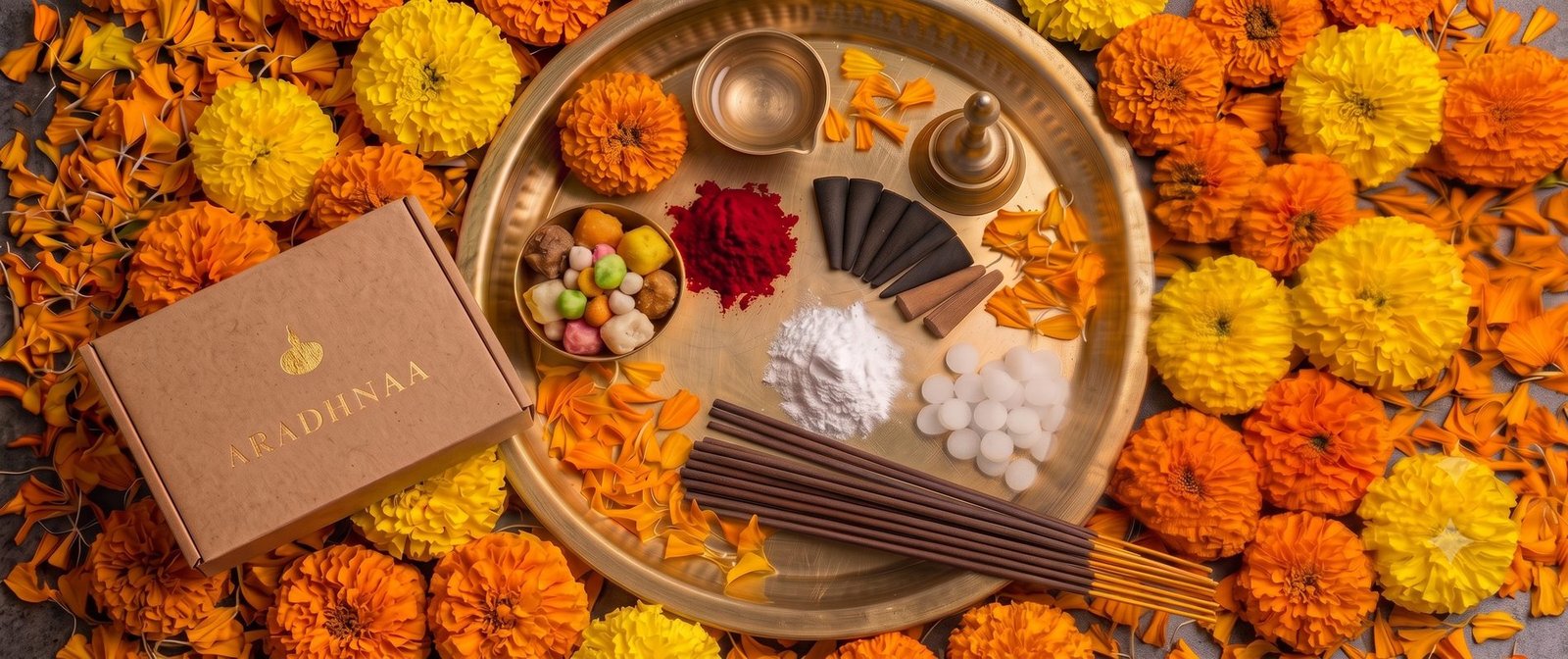 A brass pooja thali of samagri laid out among marigolds, beside an Aradhnaa box